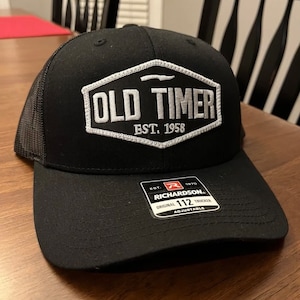 May include: Black trucker hat with a white embroidered patch that says "OLD TIMER EST. 1958". The hat has a black mesh back and a black adjustable strap with a white label that says "RICHARDSON. ORIGINAL 112 TRUCKER ADJUSTABLE".
