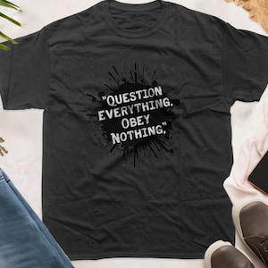 May include: Dark grey t-shirt featuring the quote "QUESTION EVERYTHING, OBEY NOTHING" in white text on a black ink splatter design. The shirt is displayed on a white surface with shoes, sunglasses, and a smartphone.