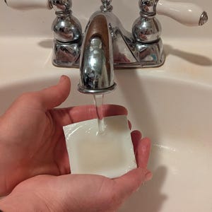 May include: A chrome faucet with white and chrome handles is running water onto a white square object held in two hands. The faucet is mounted on a white sink.