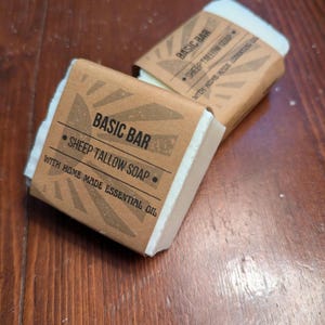 May include: Two rectangular bars of soap wrapped in brown paper. The paper is printed with the words "BASIC BAR", "SHEEP TALLOW SOAP", and "WITH HOME MADE ESSENTIAL OIL". The soap is white and appears to be handmade.