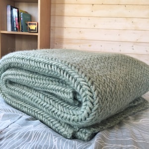 Peut inclure: Une couverture en tricot douce de couleur vert sauge pliée sur un lit avec une housse de couette à motifs blancs et bleus.