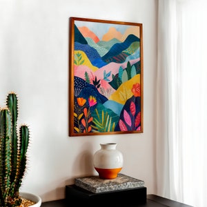 May include: A framed art print depicting a vibrant abstract landscape with hills in blue, pink, yellow, and green. The artwork is in a wooden frame and hangs on a white wall. A white and orange vase sits on a stone block.