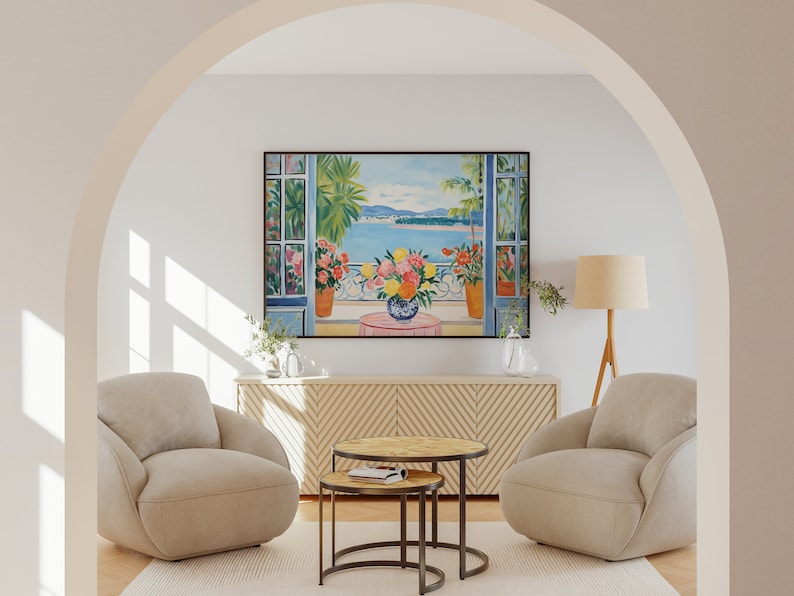 K&ouml;nnte beinhalten: Zwei beige Sessel mit einem Holz-Couchtisch und einem Beistelltisch vor einem beige Schrank mit einem Gem&auml;lde einer Meereslandschaft mit einem Balkon und Blumen in T&ouml;pfen. Das Gem&auml;lde ist in Schwarz gerahmt.