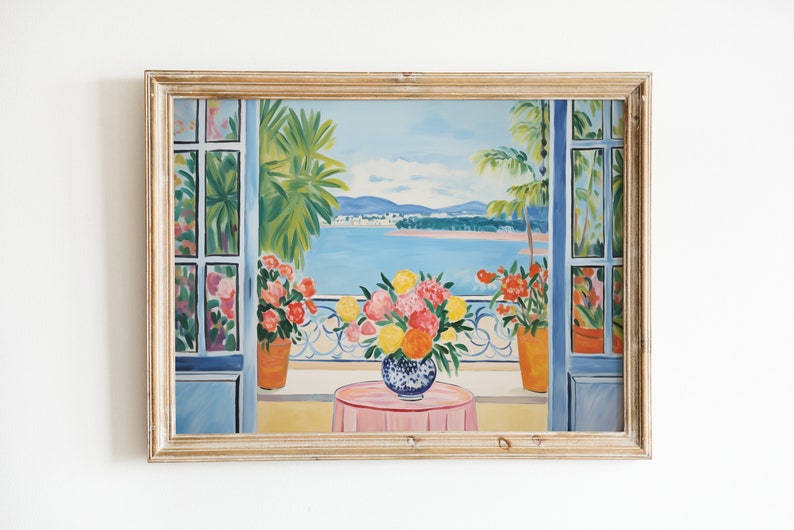 K&ouml;nnte beinhalten: Ein gerahmtes Gem&auml;lde, das einen Blick aus einem Fenster mit einem Balkon zeigt. Die Szene zeigt eine blaue Wasserfl&auml;che mit einer Stadt in der Ferne, Palmen und eine Vase mit bunten Blumen auf einer rosa Tischdecke.