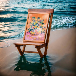 May include: A wooden folding chair with a painting of a bowl of lemons and flowers on the back. The chair is sitting on a sandy beach with blue water in the background.