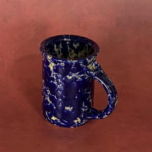 May include: A cylindrical ceramic mug featuring a deep navy blue base color with a speckled, marbled pattern of light yellow and white accents throughout the exterior and interior surfaces, complete with a curved handle on the right side.