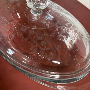 Könnte beinhalten: Eine ovale Auflaufform aus klarem Glas mit passendem Deckel. Der Deckel ist mit einem geprägten Blumenmuster aus Stielen, Blättern und kleinen Blüten verziert und wird von einem klaren Glasknauf in Blütenform gekrönt.