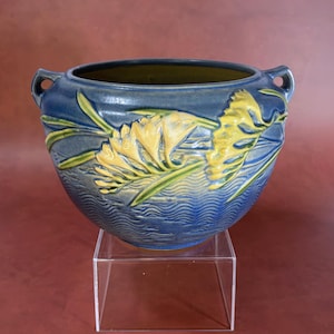 Peut inclure: Un bol en céramique rond avec deux petites anses, présentant une glaçure bleu profond et un motif en relief de fleurs jaunes et de feuilles vertes. La partie inférieure du bol affiche un design texturé à lignes ondulées, l'intérieur est vert olive.