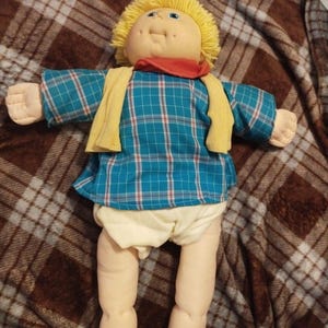 Può includere: Una bambola vintage con un viso morbido e rotondo, occhi blu e capelli di filo giallo. Indossa una camicia a quadri blu, una sciarpa gialla e un fazzoletto rosso. La bambola ha pantaloncini color crema ed è sdraiata su una coperta a quadri marrone.