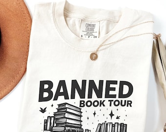 Puede incluir: Camiseta color crema con la frase "BANNED BOOK TOUR" en letras negras. El diseño incluye una pila de libros y una estantería. Un collar dorado con un colgante descansa sobre la camiseta.