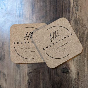 May include: Two square cork coasters with a black circle design. The circle contains the text "HK" in cursive lettering and the words "Engravings" and "Laser Engraving" in all caps. The text "Estd. 2024" is also included in the circle.