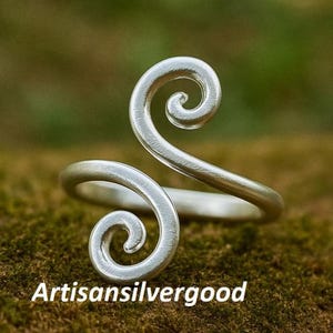 May include: A silver ring with a distinctive, swirling pattern. The ring has two opposing spiral shapes, forming an open, adjustable design. It is set against a natural, mossy background. The text "Artisansilvergood" is visible.