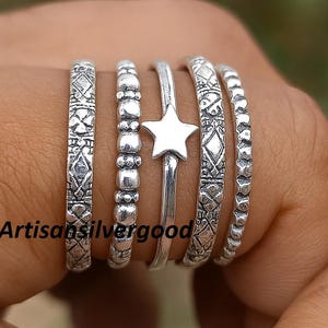 Puede incluir: Un conjunto de anillos de plata con varios diseños, incluyendo un anillo en forma de estrella y anillos con patrones geométricos. Los anillos se muestran en una mano, mostrando sus intrincados detalles y texturas. El texto "Artisansilvergood" es visible.