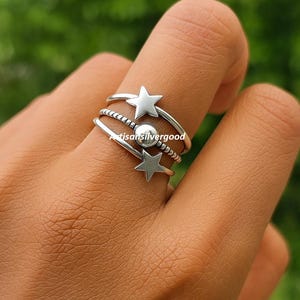 Peut inclure: Ensemble de bagues en argent avec des motifs d'étoiles et de perles. Les bagues sont empilées sur un doigt, mettant en valeur les différentes textures et formes. Les bagues comprennent une étoile, une bande perlée et une bande lisse avec une petite boule argentée.