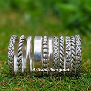 以下が含まれることがあります： 編み込み、模様、滑らかなバンドなど、さまざまなデザインのシルバーリングのコレクション。 リングは緑の草の上に表示され、「Artisansilvergood」のテキストが表示されます。