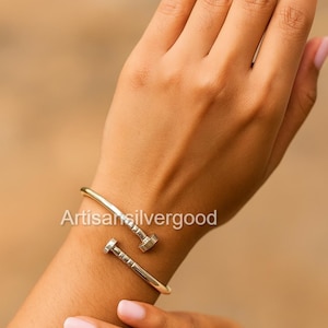 Puede incluir: Pulsera plateada con forma de clavo. La pulsera presenta dos diseños de cabeza de clavo. El texto "Artisan silvergood" es visible. La pulsera se lleva en una muñeca con un fondo neutro.