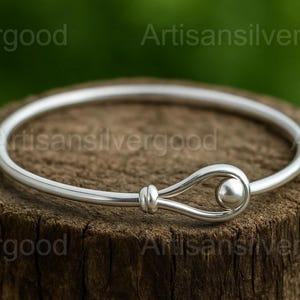 May include: A silver bangle bracelet with a hook and eye clasp. The bracelet is round and has a smooth, polished finish. The clasp features a loop design with a small ball at the end. The bracelet is displayed on a wooden surface.