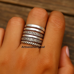 May include: A stack of silver rings with various designs, including smooth bands, patterned bands, and a twisted rope-like band. The rings are worn on a finger. The text "Artisansilvergood" is visible.
