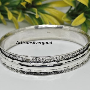 Könnte beinhalten: Ein silberfarbenes Armreif-Armband mit einem eingravierten Blumenmuster. Das Armband hat eine Reihe von strukturierten Bändern und den Text "Artisansilvergood". Der Schmuck wird vor einem Hintergrund aus grünem und gelbem Laub gezeigt.