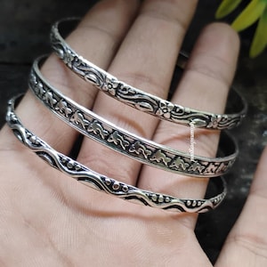 May include: Three silver bangles with detailed floral and geometric patterns. The bangles are stacked, displaying the intricate designs. The metal has a slightly oxidized finish, enhancing the details.