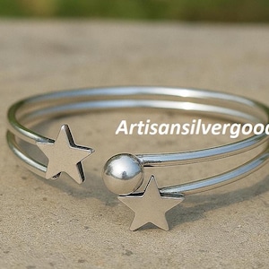 Könnte beinhalten: Ein silberfarbenes Armband mit einem himmlischen Thema. Das offene Manschettendesign hat zwei sternförmige Akzente und eine kleine Kugel. Der Text "Artisansilvergood" ist auf dem Armband zu sehen.