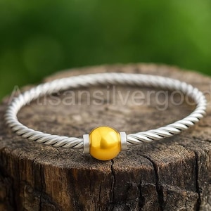 May include: A silver-colored twisted metal bracelet with a bright yellow sphere in the center. The sphere is held in place by two silver-colored metal cylinders. The bracelet is resting on a wooden surface.