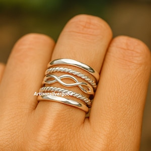 Può includere: Una mano con una pila di anelli d'argento. Gli anelli includono una fascia liscia, un design a corda intrecciata, un simbolo dell'infinito e un altro design a corda intrecciata. Gli anelli sono di Artisansilvergood.