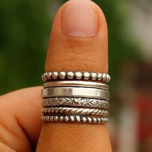 Puede incluir: Una colección de anillos de plata de varios diseños apilados en un dedo. Los anillos incluyen estilos con cuentas, lisos, estampados y de cuerda trenzada. El fondo es borroso y verde.