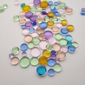 May include: A collection of colorful, translucent glass pebbles in various sizes. The pebbles are in shades of blue, green, yellow, purple, and pink. The round glass stones are scattered on a white surface.