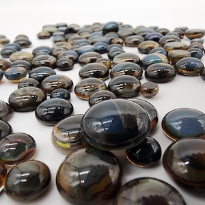 May include: A collection of smooth, iridescent glass pebbles in various sizes. The pebbles have a glossy finish and display a range of colors, including blues, browns, and greens. The pebbles are scattered across a white surface.