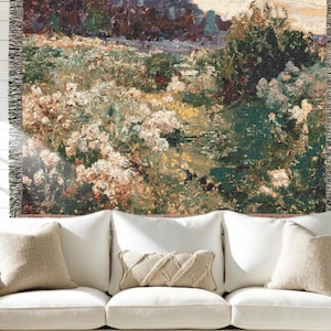 May include: A woven tapestry throw blanket featuring a landscape scene with white and yellow wildflowers, deep green foliage, and dark brown trees. The rectangular textile has a fringed edge on the left and right sides with a textured weave.