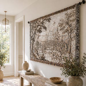 May include: A rectangular woven wall tapestry featuring a detailed monochromatic landscape scene with trees, a building, and a crowd of people. The tapestry has a fringed border and is suspended from a black metal rod mounted on a wall.