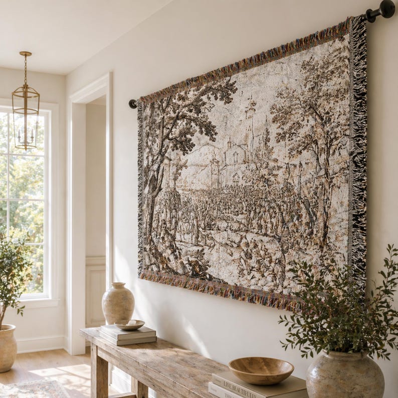 May include: A rectangular woven wall tapestry featuring a detailed monochromatic landscape scene with trees, a building, and a crowd of people. The tapestry has a fringed border and is suspended from a black metal rod mounted on a wall.