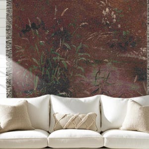 May include: A woven throw blanket with a nature-inspired design, featuring a landscape of tall green grasses and foliage against a reddish-brown background. The blanket has a fringed edge and is draped over a white sofa with beige pillows.