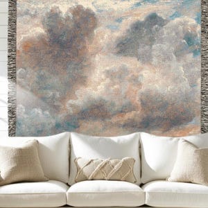 Op de afbeelding: Een geweven sprei met een wolkenluchtontwerp in beige, grijs en blauw. De deken heeft een gefranjerde rand en wordt getoond over een witte bank met beige kussens.