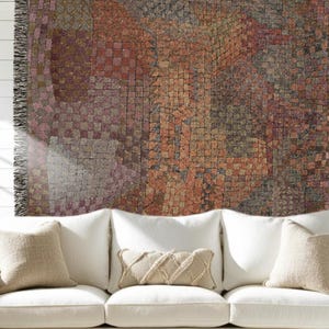 May include: A woven throw blanket featuring a mosaic-like pattern of small squares in shades of rust, brown, grey, and muted purple. The blanket has a fringed edge on the left side and is draped over a white sofa with several pillows.