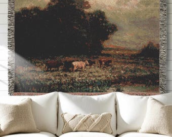 May include: A woven tapestry throw blanket featuring a landscape scene with a large dark tree cluster, a field, and several cows grazing. The blanket has a fringed edge and displays a color palette of deep greens, browns, and muted earth tones.