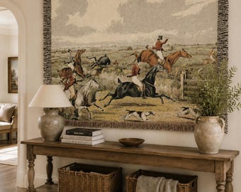 May include: A large rectangular woven wall tapestry featuring a detailed scene of a fox hunt with riders on horseback and hounds in a field. The tapestry has a fringed border and displays muted earth tones including brown, tan, and green.