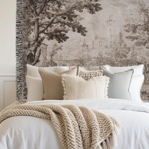 May include: A woven wall tapestry featuring a monochromatic landscape illustration of trees, buildings, and a town square. The textile has a fringed edge on the left side and is displayed above a bed with white bedding and several neutral-colored pillows.