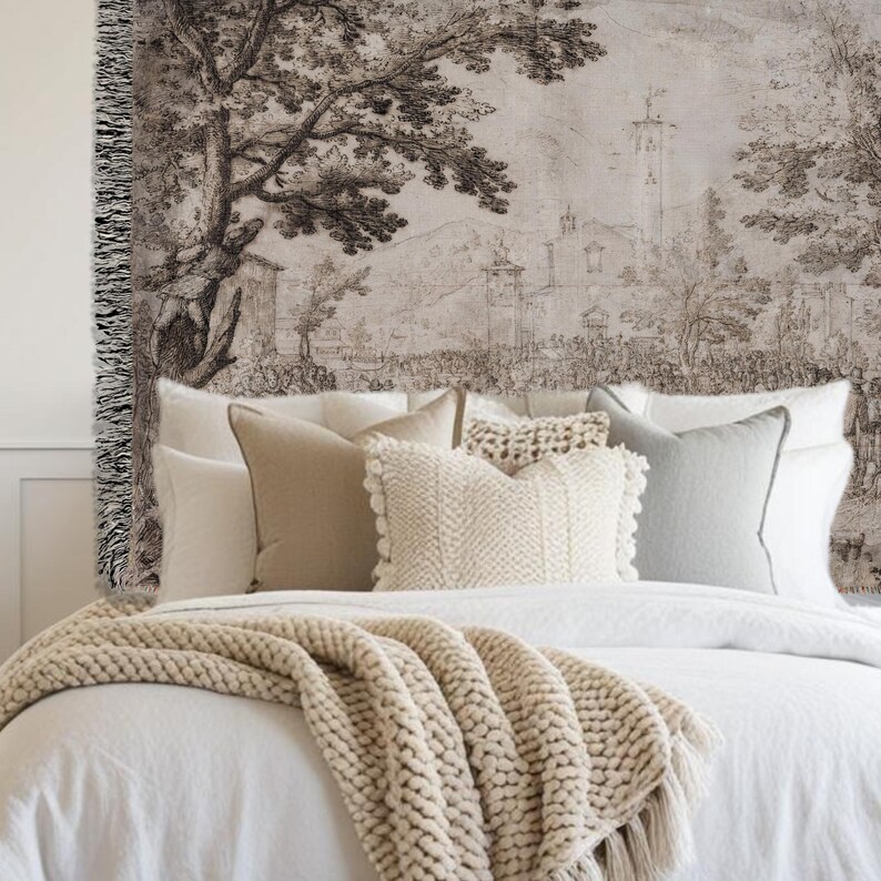 May include: A woven wall tapestry featuring a monochromatic landscape illustration of trees, buildings, and a town square. The textile has a fringed edge on the left side and is displayed above a bed with white bedding and several neutral-colored pillows.