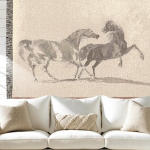 May include: A woven throw blanket featuring a sepia-toned illustration of two horses. One horse is rearing up on its hind legs, while the other stands nearby. The blanket has a fringed edge and is displayed over a white couch with beige pillows.