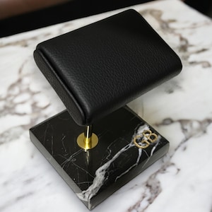 May include: A black leather watch stand with a gold metal base and a black marble base with the initials "GB" engraved in gold.