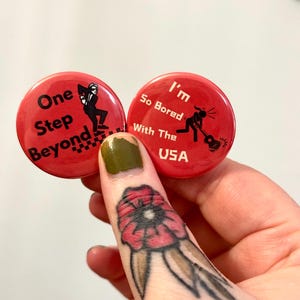 May include: Two circular red pinback buttons. The left button features a black silhouette of a person in a suit and hat with the text "One Step Beyond" and a checkered pattern. The right button shows a black silhouette playing a guitar with the text "I'm So Bored With The USA".