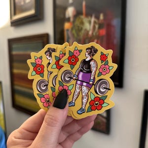 May include: A die-cut sticker featuring a person with tattoos lifting a barbell. The person wears a black tank top and purple shorts. The design includes red and pink flowers on a yellow background. The text "mellowtattoos" is printed on the side.