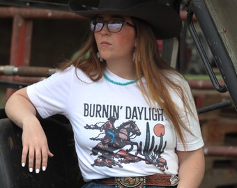Może przedstawiać: Kobieta w białej koszulce z napisem "Burnin' Daylight" i grafiką kowboja na koniu. Ma na sobie czarny kowbojski kapelusz, niebieskie dżinsy i brązowe buty. Siedzi w czarno-białym pojeździe użytkowym.