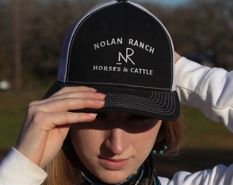 Puede incluir: Una mujer lleva una gorra de camionero negra y blanca con el texto "Nolan Ranch NR Horses & Cattle" bordado en la parte delantera. También lleva un pañuelo de paisley azul turquesa alrededor del cuello.