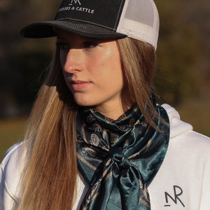 Może przedstawiać: Kobieta w białej bluzie z kapturem z czarno-białym logo "NR", czarną i białą czapką trucker z napisem "Nolan Ranch Horses & Cattle" i ciemnozielonym bandaną paisley zawiązaną wok&oacute;ł szyi.