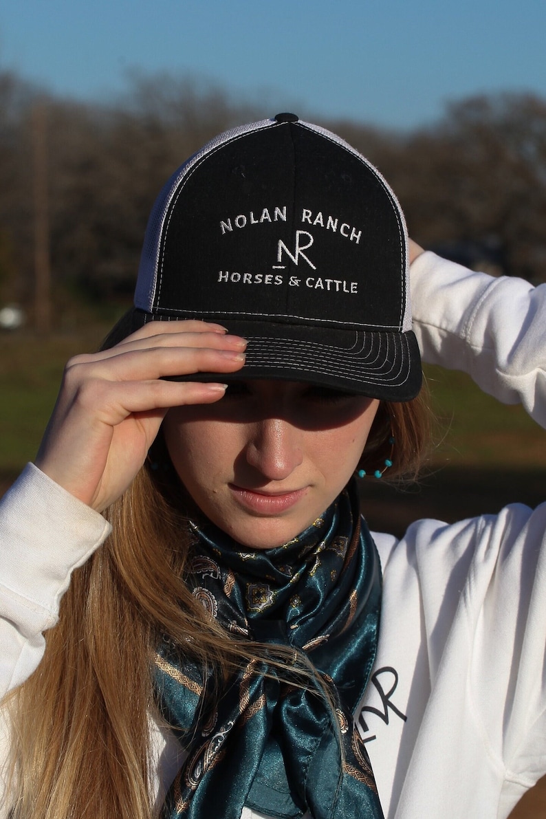 Może przedstawiać: Kobieta w czarno-białej czapce trucker z haftem "Nolan Ranch NR Horses & Cattle" z przodu. Nosi r&oacute;wnież turkusowy bandanę w paisley wok&oacute;ł szyi.