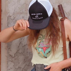 Może przedstawiać: Czarno-biała czapka trucker z siateczką z tyłu. Na czapce jest wyszyty napis "Nolan Ranch NR Horses & Cattle".