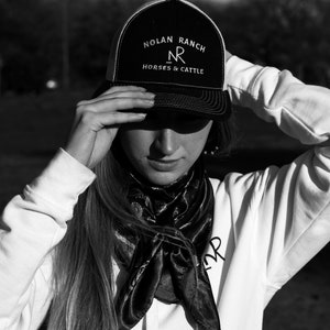 Może przedstawiać: Osoba w czarno-białej czapce trucker z napisem "Nolan Ranch" i "Horses & Cattle" z przodu. Osoba nosi r&oacute;wnież białą bluzę z kapturem i czarną chustkę.